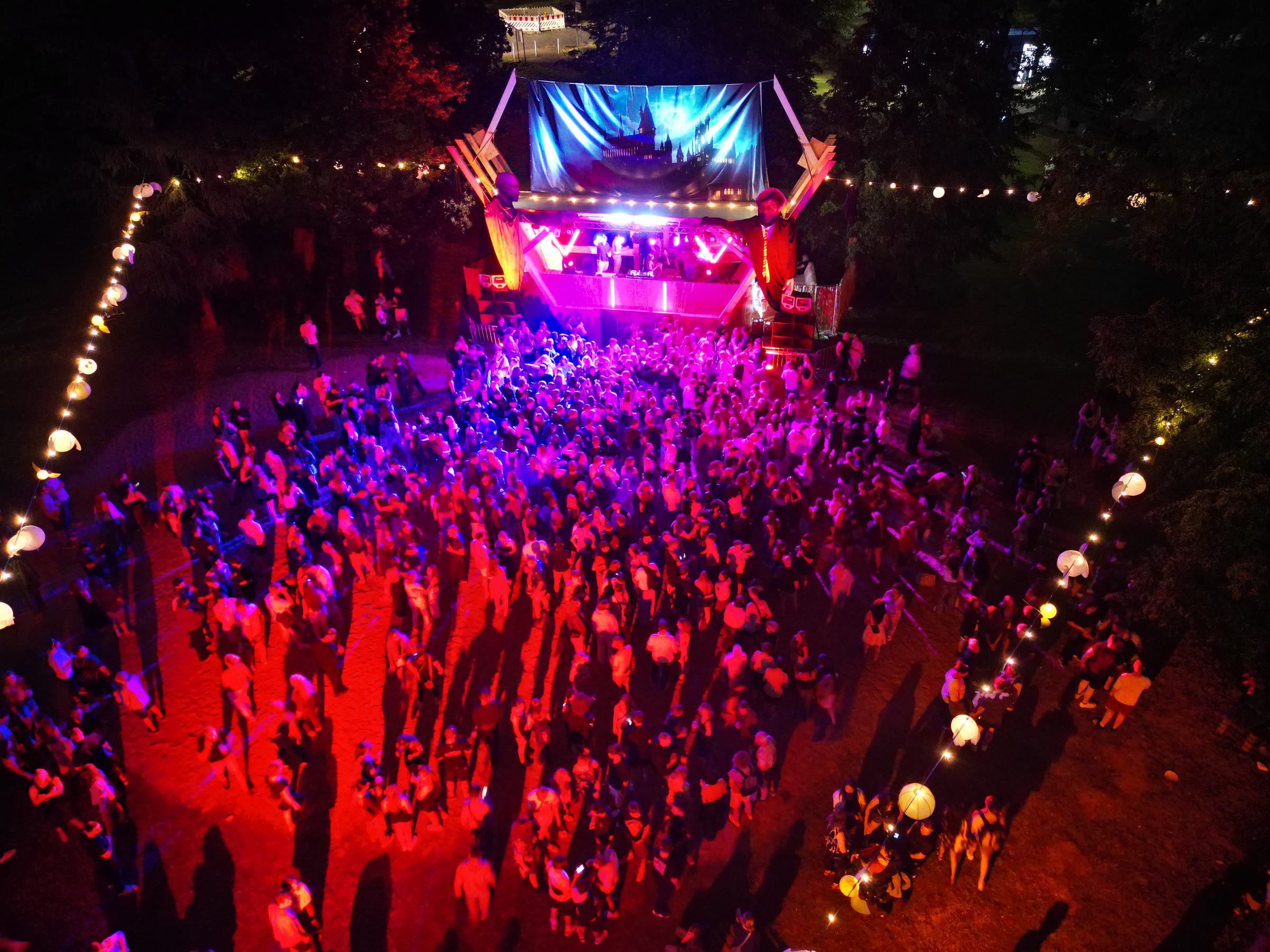Drohnenaufnahme des Brunnenfests bei Nacht: die leuchtende Bühne und eine tanzende Menge, gerahmt von Lichterketten
